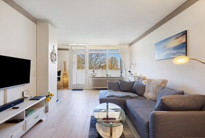 En moderne stue med et fladskærms-tv, et glasbord, en grå sofa og et stort vindue med gardiner.