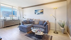 Un salon moderne avec un canapé d’angle, une table basse en verre, un lampadaire et une vue sur les immeubles à travers de grandes fenêtres.