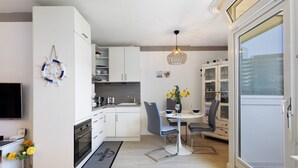 Une cuisine moderne avec des meubles blancs, un coin repas comprenant une table et des chaises, et une grande fenêtre donnant sur des immeubles.