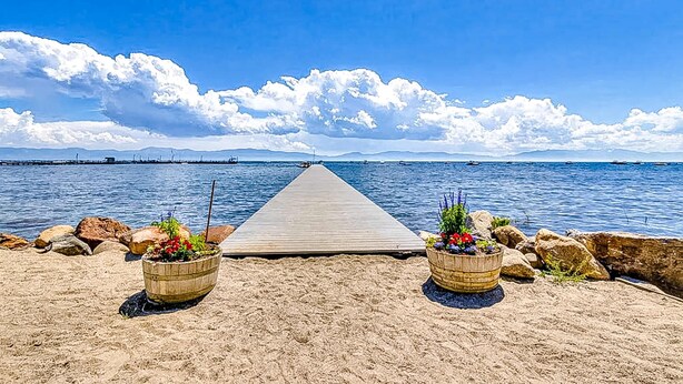 Un muelle de madera que se extiende hacia un mar tranquilo bajo un cielo despejado, rodeado de rocas y dos macetas con flores.