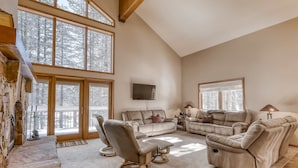 A spacious living room with a stone fireplace, large windows, a beige sofa, and a flat-screen TV. - Three-story lakefront home with lake views, wood fireplace, high-speed WiFi, W&D (Soda Springs)