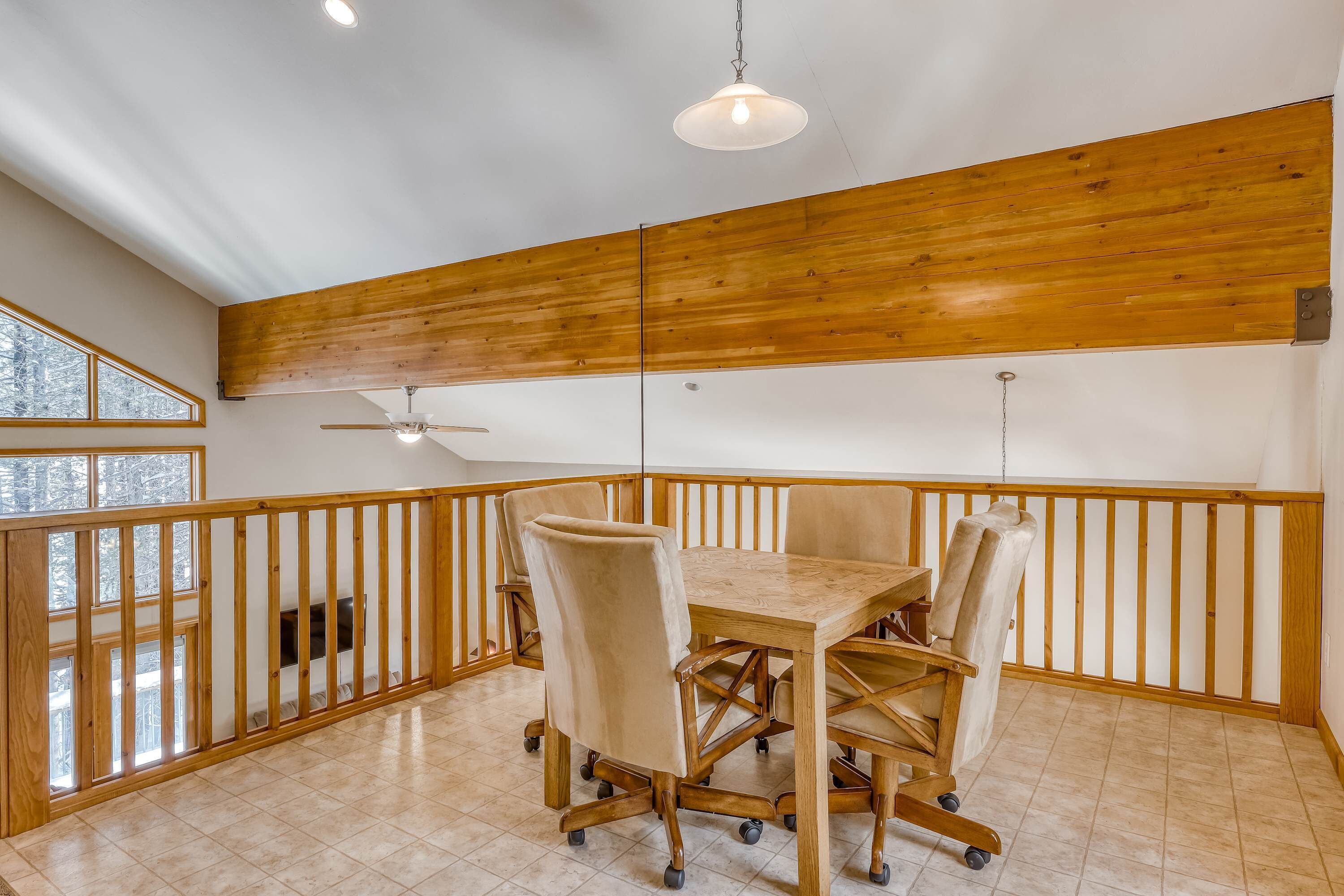 A room with a wooden dining table, chairs, a ceiling fan, and a view of trees through large windows.