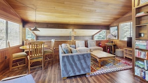 A cozy cabin interior with a wooden ceiling, a sofa, chairs, a small table, a bookshelf, and a window with blinds. - Spacious Northstar Lake Retreat – Perfect for Families & Groups ️ (Truckee)