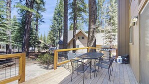 A wooden deck with a table and chairs, surrounded by tall trees, and a building in the background. - Spacious Northstar Lake Retreat – Perfect for Families & Groups ️ (Truckee)