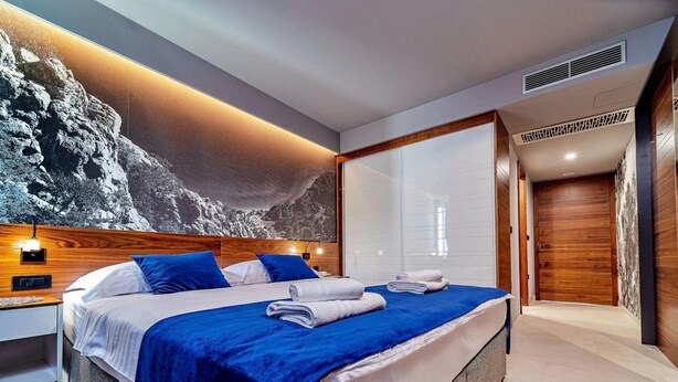 A modern bedroom with a large bed, a nightstand, a wall-mounted light, and a view of a mountainous landscape. - Stay With Pool in Promajna (Baska Voda)