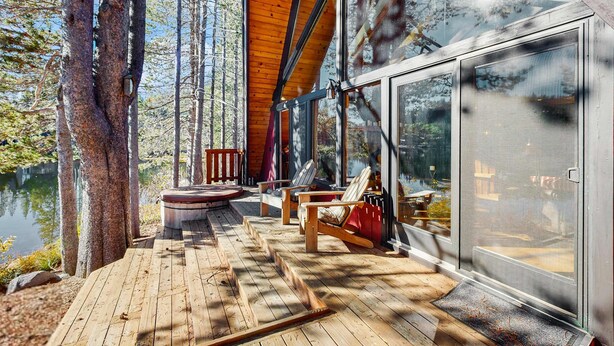 Una terraza de madera con una sala acristalada, sillas de madera y vistas a un lago.