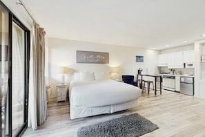 A bedroom with a large bed, a bedside table, a lamp, and a kitchen area with cabinets and appliances.