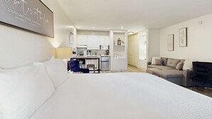 A modern hotel room with a large bed, a kitchenette, a sofa, and a dining table.