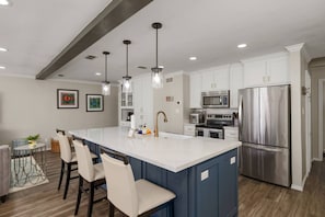 A modern kitchen with white cabinets, stainless steel appliances, a large island with bar stools, pendant lighting, and a dining area with a table and chairs. - Scallop Waterfront Cottage @ LIV `Gateway to Mars` (Port Isabel)