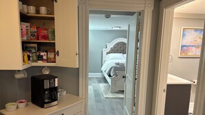 Une cuisine équipée d’une machine à café, d’un placard contenant des provisions et d’une porte donnant sur une chambre avec un lit.