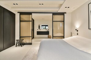 Een moderne slaapkamer met een groot bed, een klein krukje, een kaptafel met spiegel, en een schuifdeur die leidt naar een andere kamer.