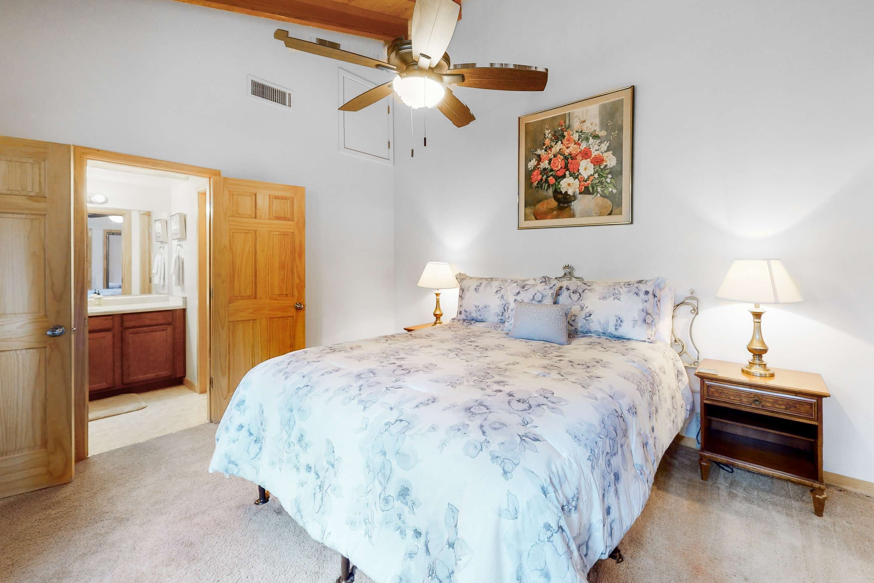 A bedroom with a bed, two bedside lamps, a ceiling fan, a painting, and a wooden door.