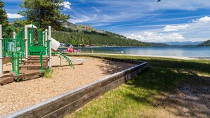 A playground with climbing structures and a slide, a wooden deck, and a lake with boats in the background. - Spacious home with private hot tub & game room - near skiing, hiking, biking (Truckee)