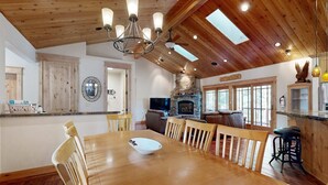 A wooden dining table with chairs, a chandelier, a stone fireplace, and a television in a well-lit room with wooden ceilings and large windows. - Immaculate Dog-Friendly Home with Private Hot Tub, Billiards, 2 Jacuzzi Tubs (South Lake Tahoe)