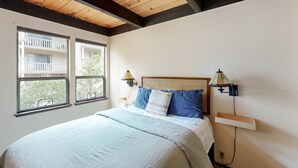 A bedroom with a bed, two windows, a nightstand, a lamp, and a shelf. - Inviting condo with balconies, fireplace, outdoor pool, sauna, & fitness center (Kings Beach)