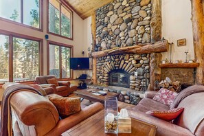 A cozy living room with leather sofas, a stone fireplace, and large windows overlooking a forest. - Modern Lux Cabin with Stunning Views, Spa, Large Kitchen and Dining, Fireplace (Carnelian Bay)