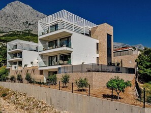 A modern two-story building with balconies, a concrete retaining wall, and a mountainous backdrop. - Stay With Rooftop Spa & Gym (Baska Voda)