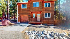A two-story wooden cabin with a front porch, garage, and a stone foundation. - Cozy, enchanting retreat with hot tub, stunning deck, & balcony - close to beach (Kings Beach)