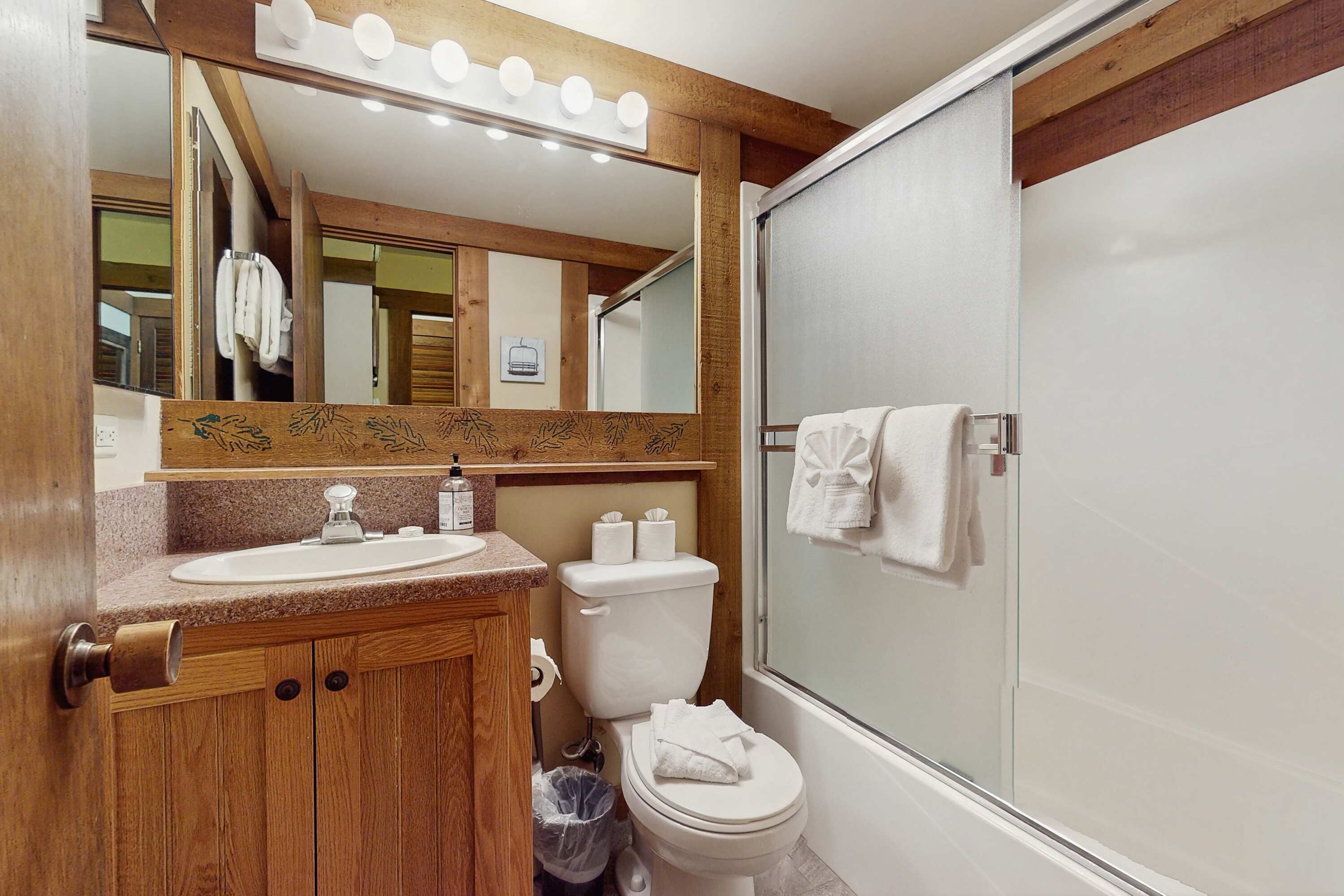Ein Badezimmer mit einem Holzbadezimmermöbel, einem Waschbecken, einer Toilette, einer Dusche mit Glastür und einem beleuchteten Spiegel.