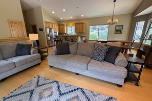 Ein geräumiges Wohnzimmer mit grauem Sofa, einem gemusterten Teppich und einer Küche im Hintergrund.