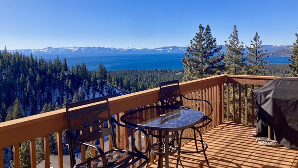 Una terraza de madera con una mesa y sillas de vidrio, con vista a un lago y montañas.