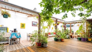En træterrasse med plantekasser, et bord dækket med dug, og en hvid væg med en billedramme.