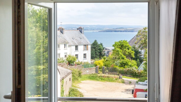 Udsigt gennem et vindue af et boligområde med huse, en grusvej og et vandområde i det fjerne.