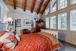 A bedroom with a large bed, wooden beams on the ceiling, a dresser, and large windows.