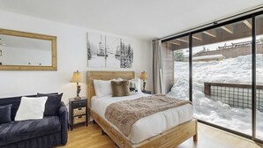 A bedroom with a bed, a sofa, a nightstand, a lamp, and a large window with a view of snow outside. - Cozy studio condo with kitchenette, deck, WiFi, & nearby activities (Truckee)