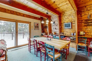 A wooden cabin interior with a dining table, chairs, a fireplace, and a large window overlooking a snowy landscape.