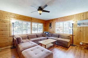 A log cabin interior with a sectional sofa, a wooden coffee table, a ceiling fan, and large windows.