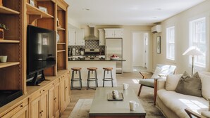 Una cocina moderna con armarios de madera, una isla central con taburetes, un comedor con sofá y mesa de centro, y un televisor en un estante de madera.