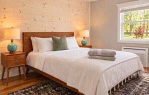 A bedroom with a wooden headboard, two bedside tables with lamps, a bed with white bedding, a window with a view of trees, and a patterned rug.