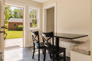 An open door leading to a patio with a table and chairs, a view of a house, and a patterned cushion on a bench.