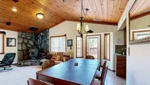 A spacious dining area with a large wooden table and chairs, a stone fireplace, and a kitchen with a stone wall and wooden ceiling. - Woodland Retreat with Private Multi-Level Deck & Backyard Creek (Truckee)