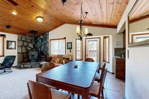 A spacious dining area with a large wooden table and chairs, a stone fireplace, and a kitchen with a stone wall and wooden ceiling. - Woodland Retreat with Private Multi-Level Deck & Backyard Creek (Truckee)