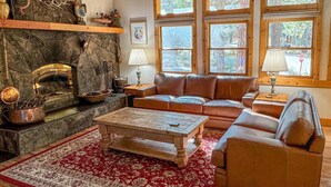 Ein gemütliches Wohnzimmer mit Kamin aus Stein, Ledersofas, einem Couchtisch und großen Fenstern.