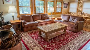 Ein gemütliches Wohnzimmer mit einem braunen Ledersofa, einem Holztisch und einem roten Teppich mit Muster. Große Fenster lassen viel natürliches Licht herein.