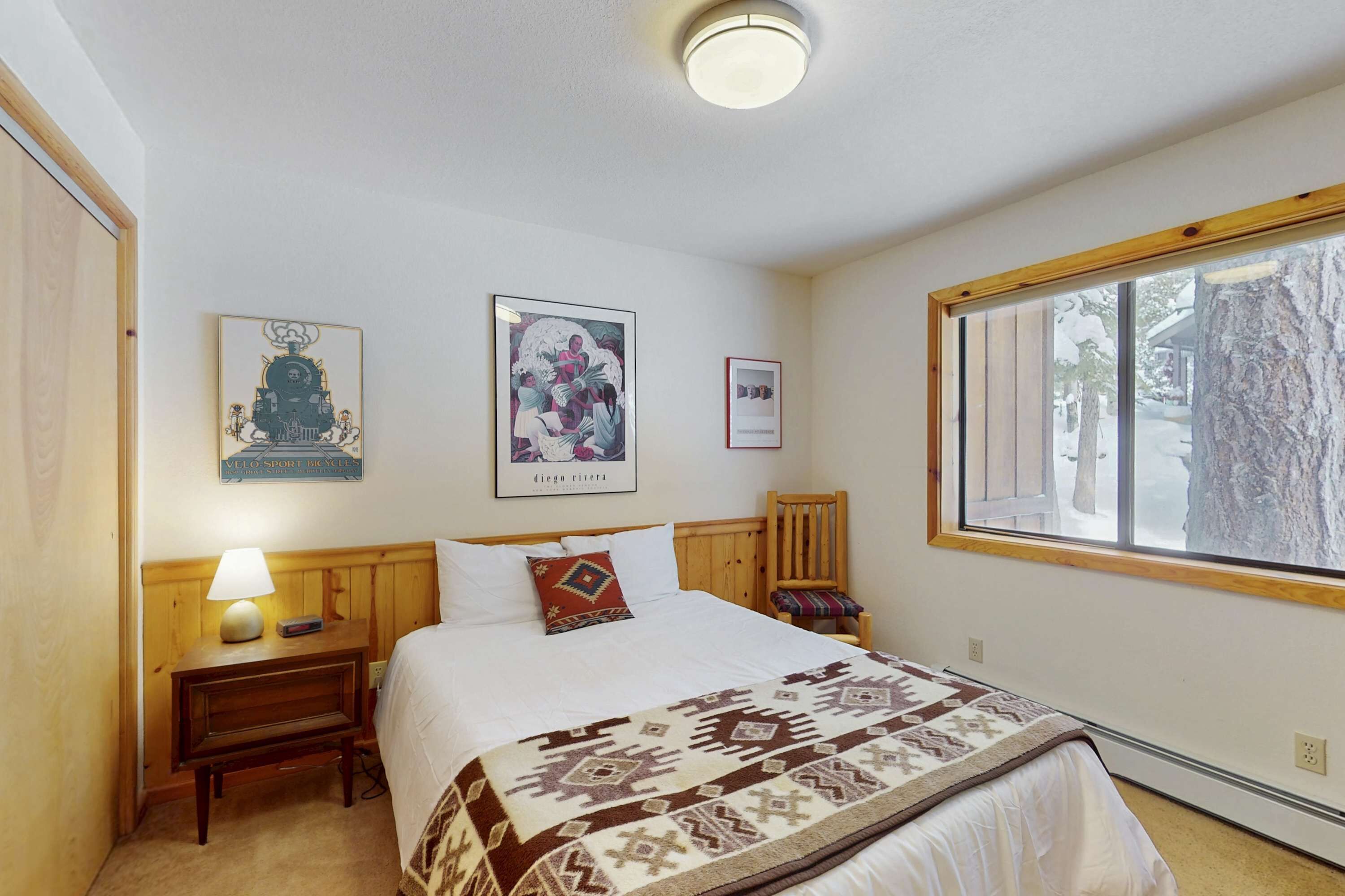 Un dormitorio con cama, mesita de noche, lámpara, silla y ventana con vistas a árboles.