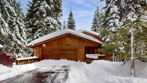 A wooden cabin with a garage, surrounded by snow-covered trees. - Rustic home with wood stove, deck, open floor plan, & nearby activities (Carnelian Bay)