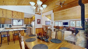 A spacious interior with wooden ceilings, a dining area with a wooden table and chairs, a kitchen with modern appliances, and a stone fireplace. - Rustic home with wood stove, deck, open floor plan, & nearby activities (Carnelian Bay)