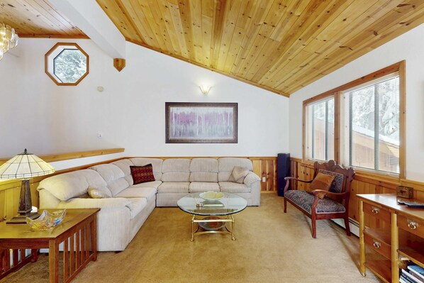 A living room with a wooden ceiling, a beige sofa, a glass coffee table, and a wooden bench. - Rustic home with wood stove, deck, open floor plan, & nearby activities (Carnelian Bay)