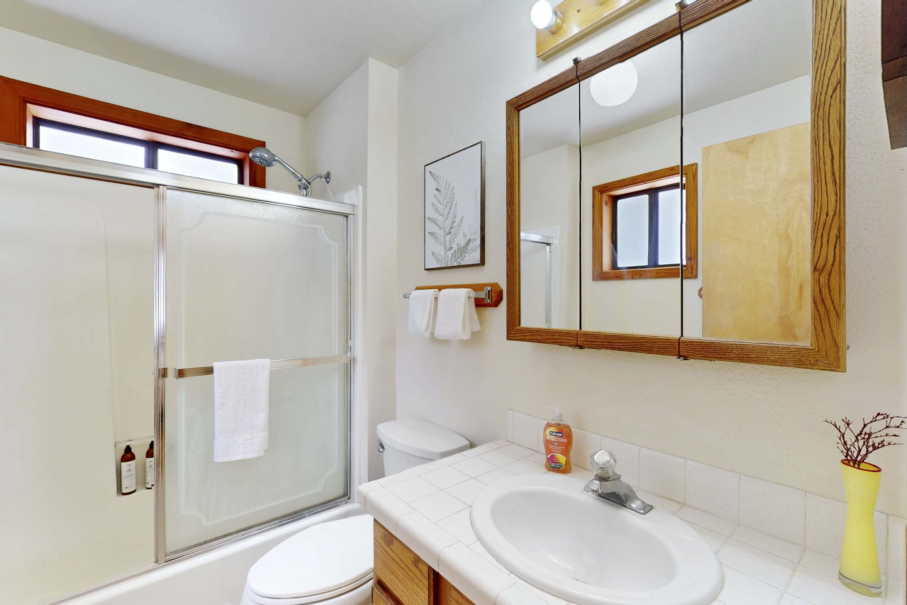 Ein Badezimmer mit Duschkabine aus Glas, einer weißen Toilette, einem Waschbecken mit Spiegel und einer Vase mit Ästen.