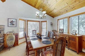 A dining area with a wooden table, chairs, a chandelier, and large windows. - Rustic home with wood stove, deck, open floor plan, & nearby activities (Carnelian Bay)