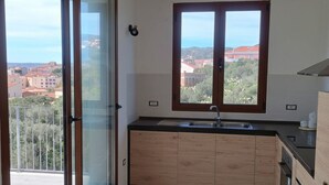A kitchen with a large window, a sliding glass door, and a view of buildings and greenery. - Maison Angelo - Sea & mountain views, panoramic terrace in Sardinia (La Maddalena)