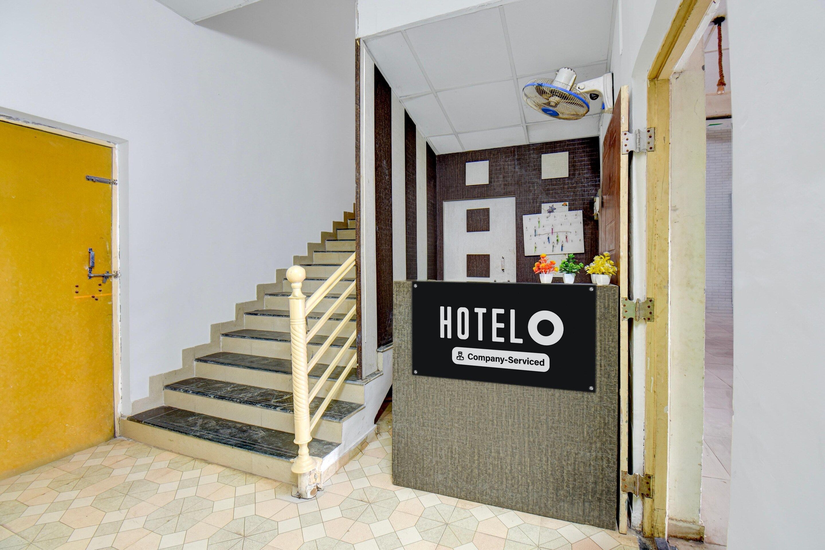 A hotel reception desk with a sign indicating it is company-serviced, a staircase with a handrail, and a yellow door to the left.