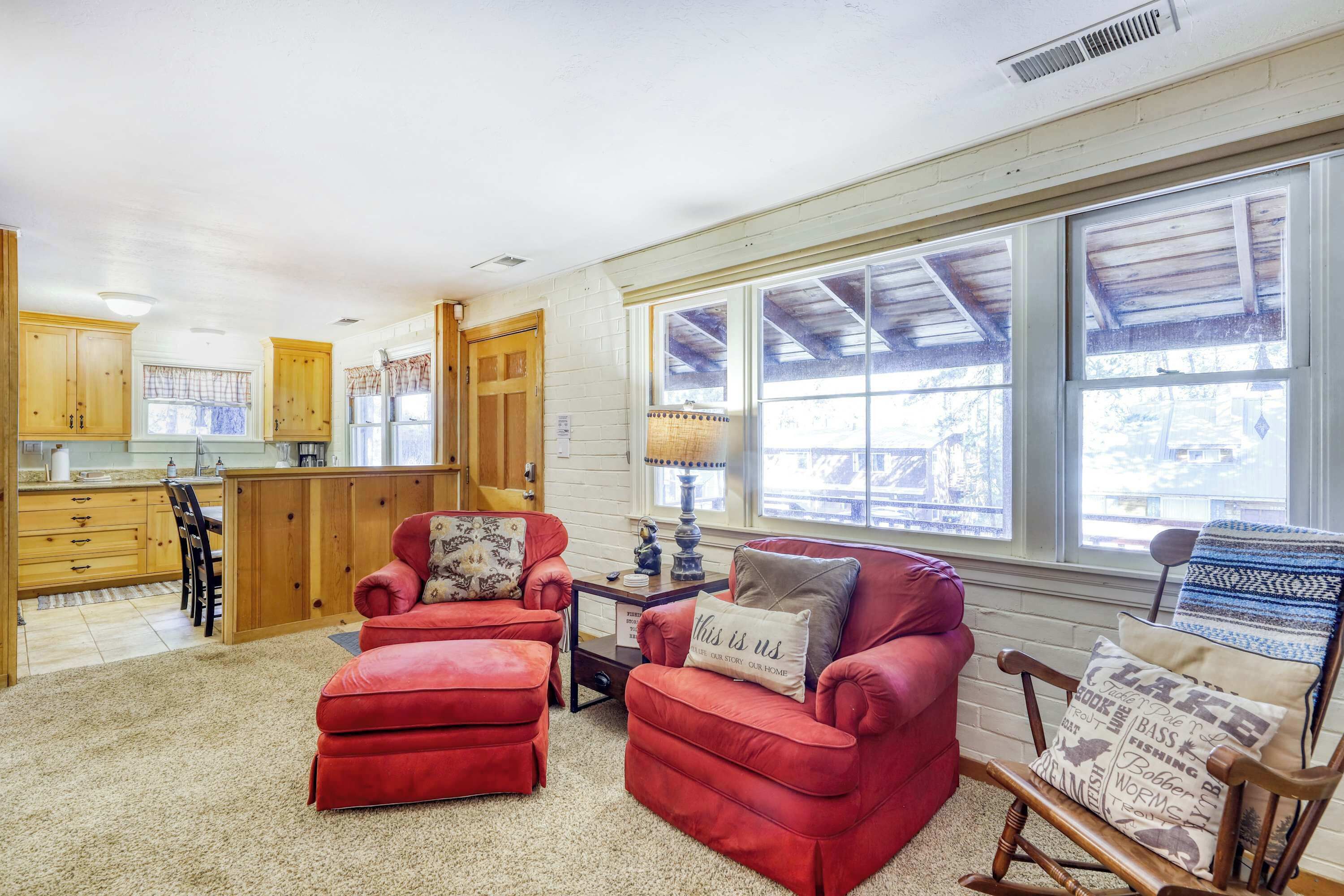 A cozy living room with red sofas and a wooden rocking chair, a kitchen with wooden cabinets, and large windows with a view of the outdoors.