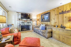 A cozy living room with a stone fireplace, a television mounted on the wall, and a wooden cabinet with decorative items. - Basecamp on Aspen (South Lake Tahoe)