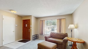 Una habitación con una puerta, una puerta de madera, una ventana con cortinas, un sofá marrón, una pequeña mesa de madera y una lámpara.