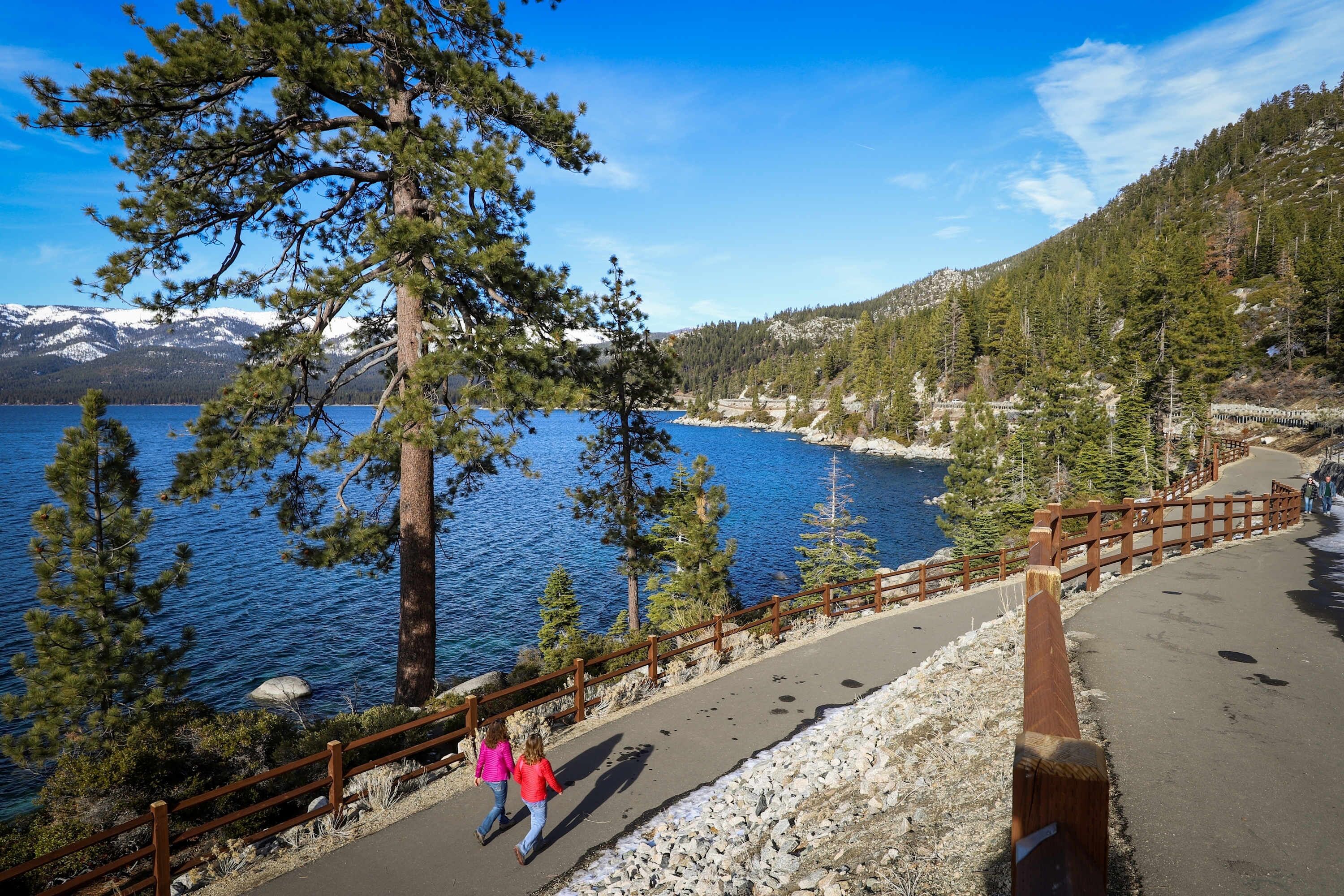 A paved walkway with a wooden railing, a lake, and a forested hillside.